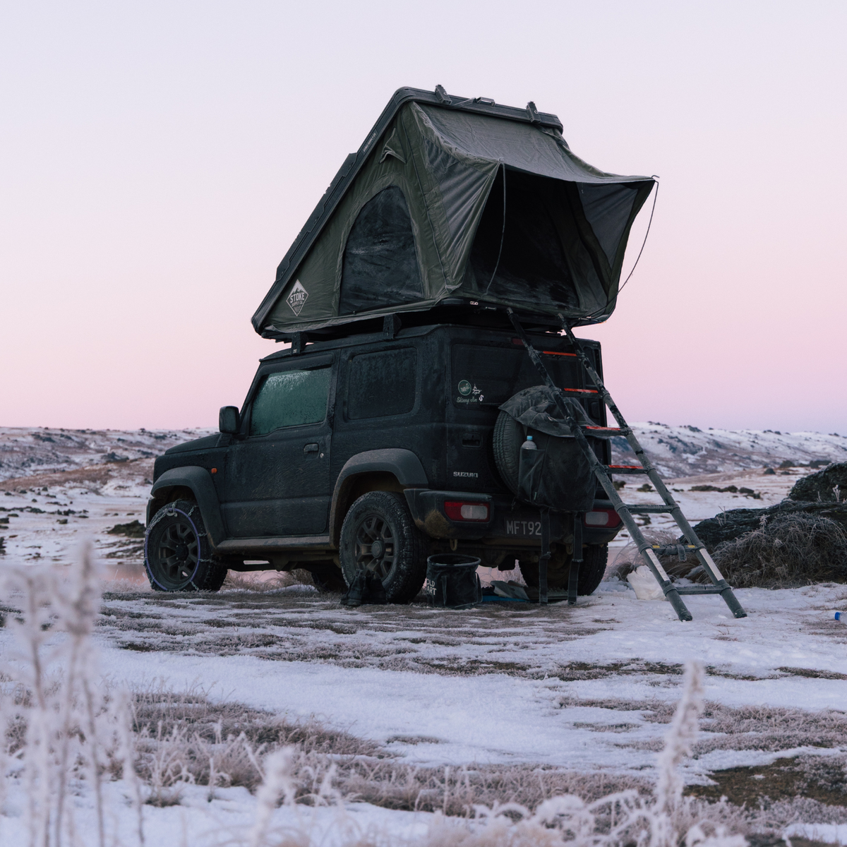 STEALTH rooftop tent open on a black Suzuki Jimny at dawn on frozen ground, telescopic ladder down and snow chains fitted