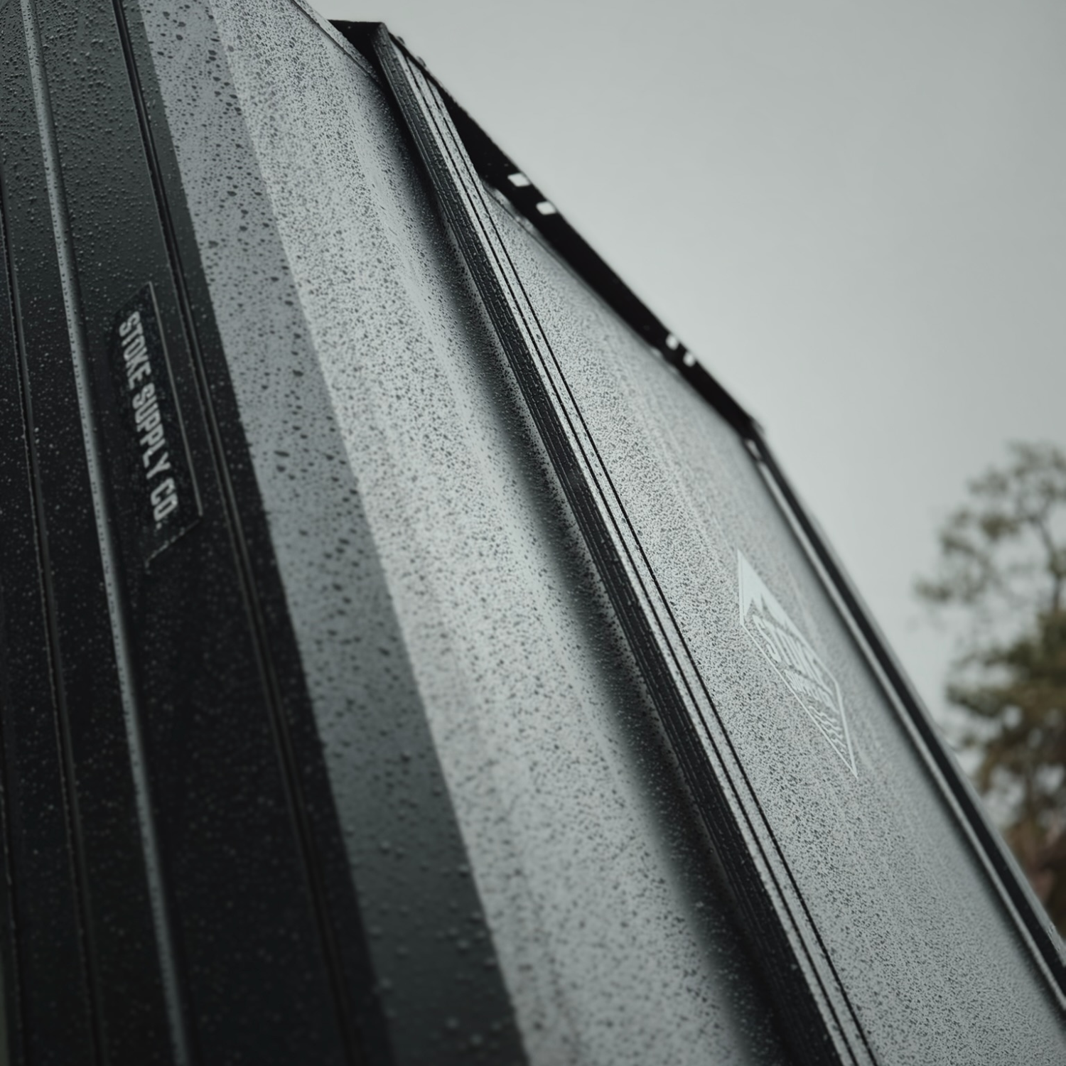 Stoke slimline crossbar mounted along the T-channel of a hard-shell rooftop tent, rain beaded on the black bar