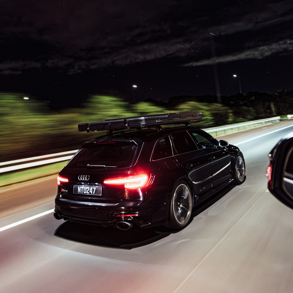 Black Audi RS4 Avant travelling at night on the motorway with a closed Stoke hard-shell rooftop tent and crossbars mounted on the roof